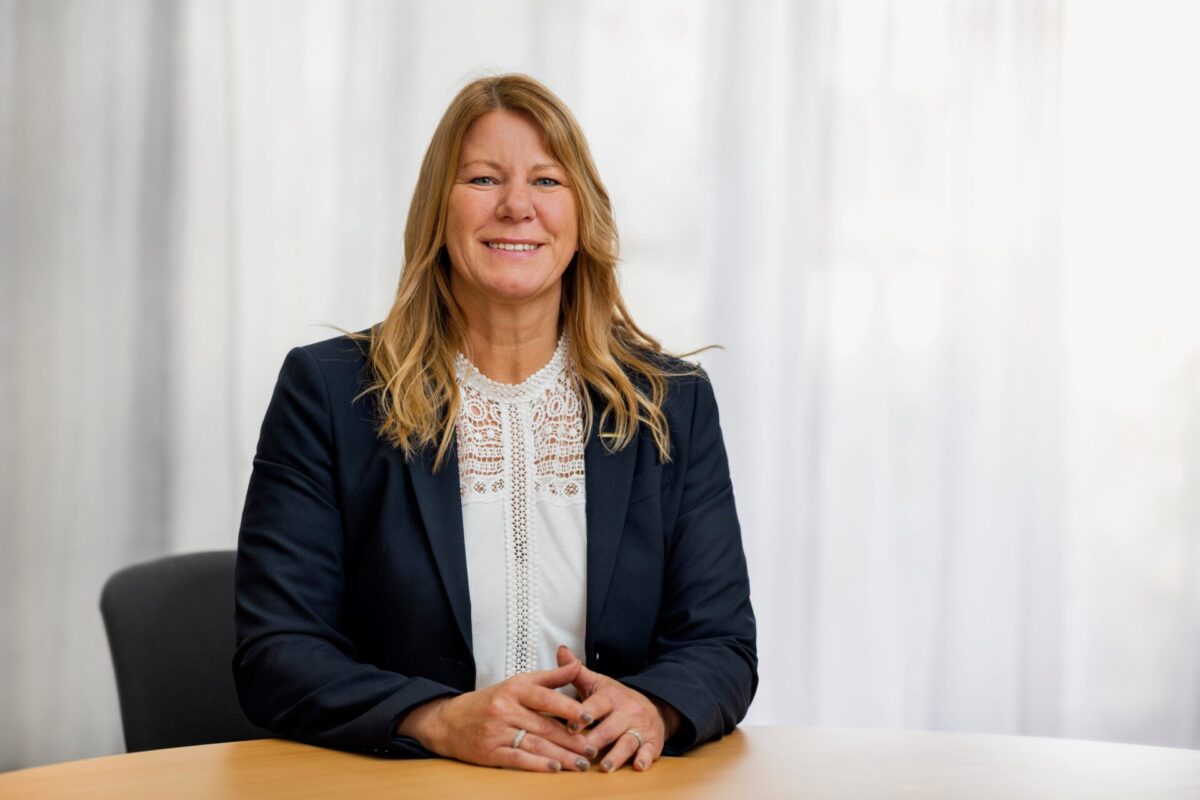 Susanne Dalskog sitter vid en skrivbord och tittar in i kameran. Hon har en mörkblå kavaj, vit skjorta och mer.